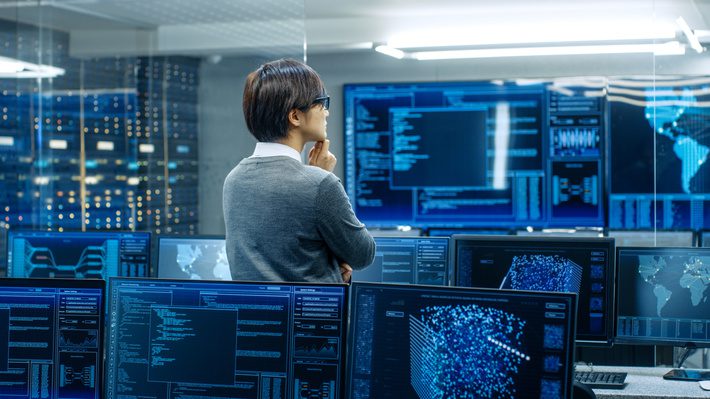 In the System Control Room Technical Operator Stands and Monitors Various Activities Showing on Multiple Displays with Graphics. Administrator Monitors Work of  Artificial Intelligence.
