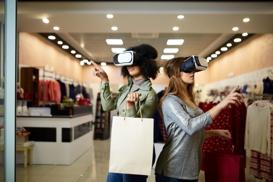 Two women in modern virtual reality headsets having expirience in shopping at lingerie store. Multiracial girls in vr glasses with bags touching and pointing interface elements in underwear shop.