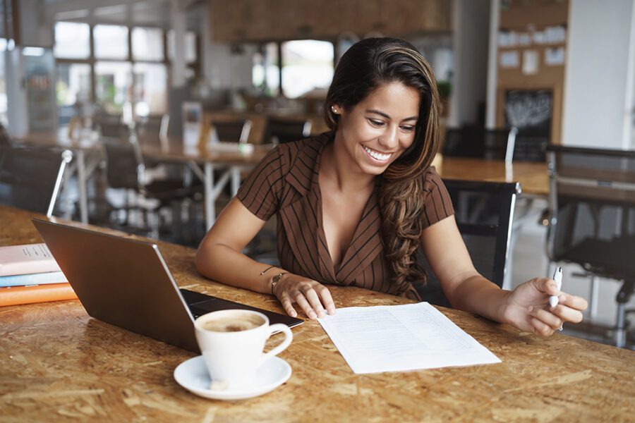 Business, gig economy concept, Charming young brazilian female entrepreneur sitting co-working cafe, reading working paper, taking notes, using laptop as working remote from office, drink cappuccino