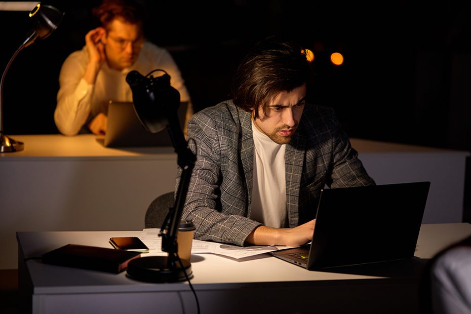 Exhausted focused young man working remotely late at night in office using computer