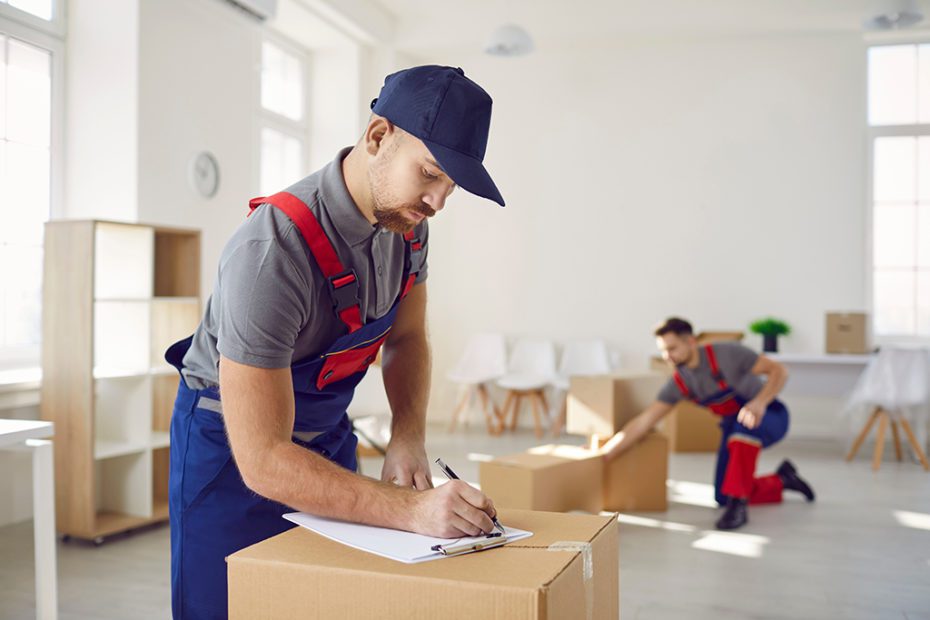 Professional male mover in overalls makes notes on clipboard while standing near cardboard boxes.