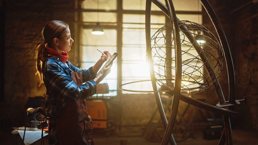 Beautiful Female Artist Sketches on a Tablet Computer Next to Brutal Metal Sculpture in Studio. Tomboy Girl Wears Checkered Shirt and Apron. Contemporary Fabricator Creating Abstract Steel Art.