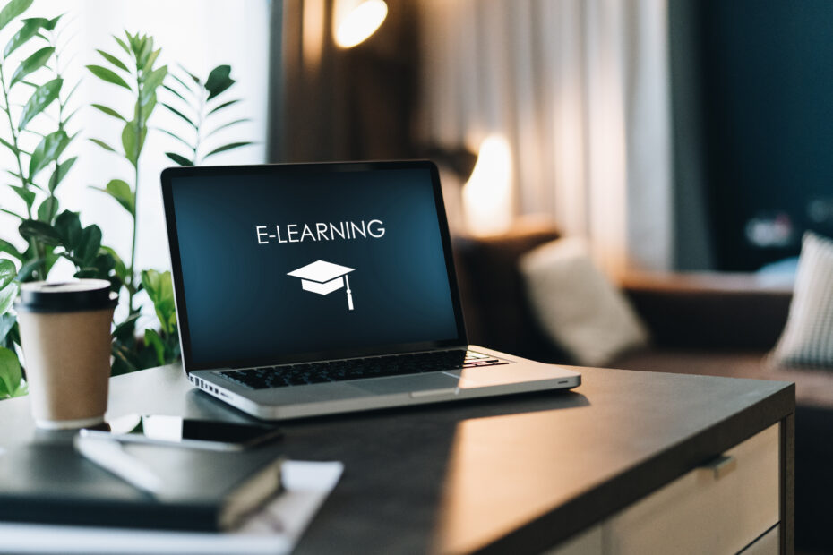 Close up of laptop with inscription on screen e-learning and image of square academic cap and smartphone on table in empty room.