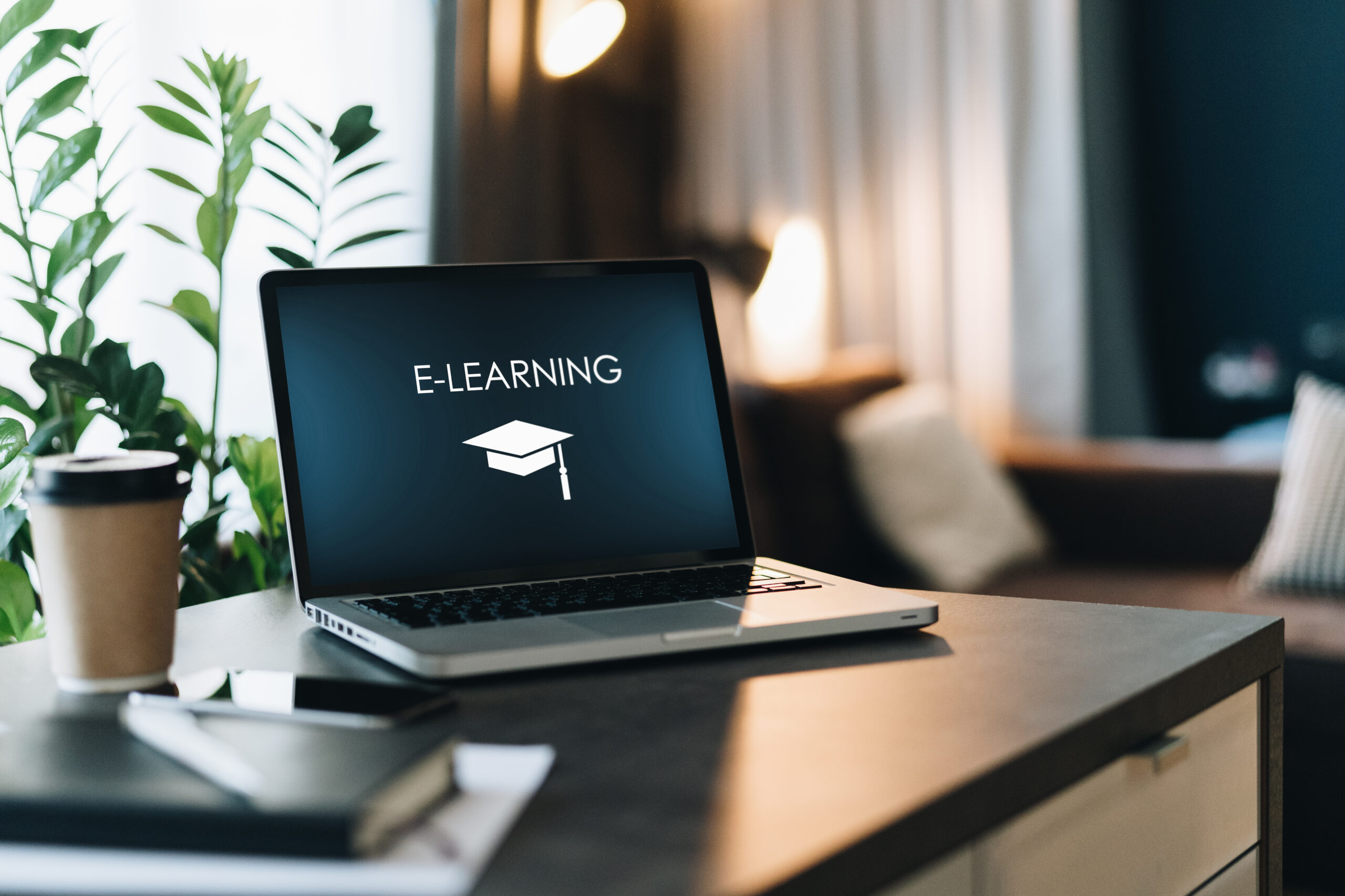 Close up of laptop with inscription on screen e-learning and image of square academic cap and smartphone on table in empty room.